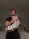 A performer in a dim bedroom hugs a long chain of banded cash bundles toward a low phone