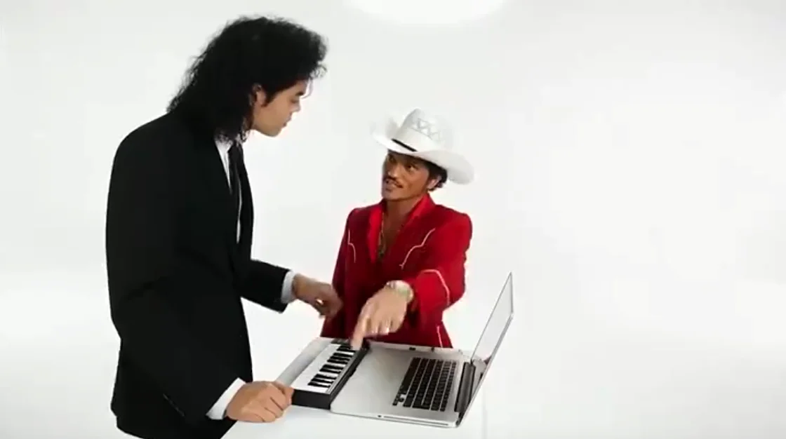 A man with long curly hair in a black suit at a compact keyboard while a man in a red western jacket and white cowboy hat reaches over to play it in a white studio