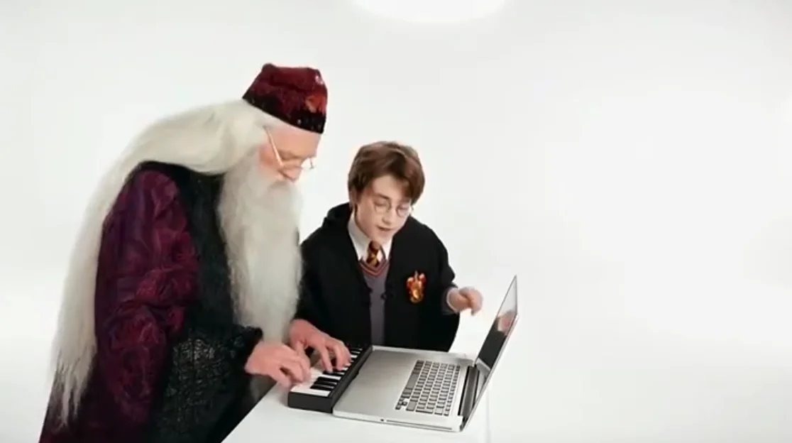 A white-bearded wizard in a velvet robe and hat playing a compact keyboard next to a young student in round glasses and a school robe in a white studio
