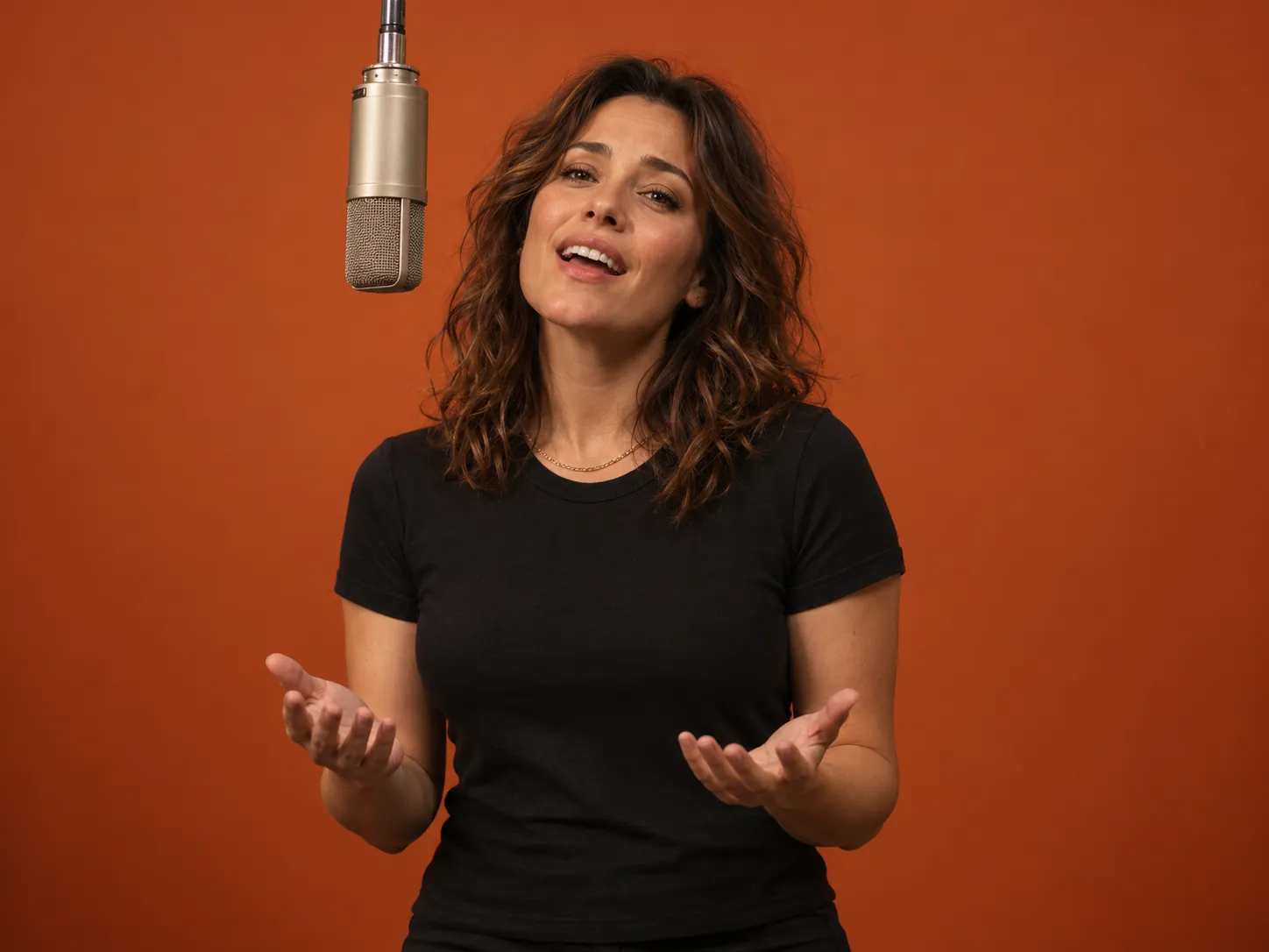 A woman in a black T-shirt and gold chain sings with open palms at a studio microphone in front of an orange wall, a Bécane Yamê AI trend serenade