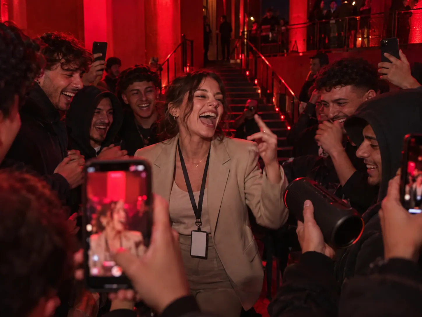 A young woman in a beige office blazer and work lanyard laughing as she freestyles at a red-lit entrance while the crowd around her films on their phones