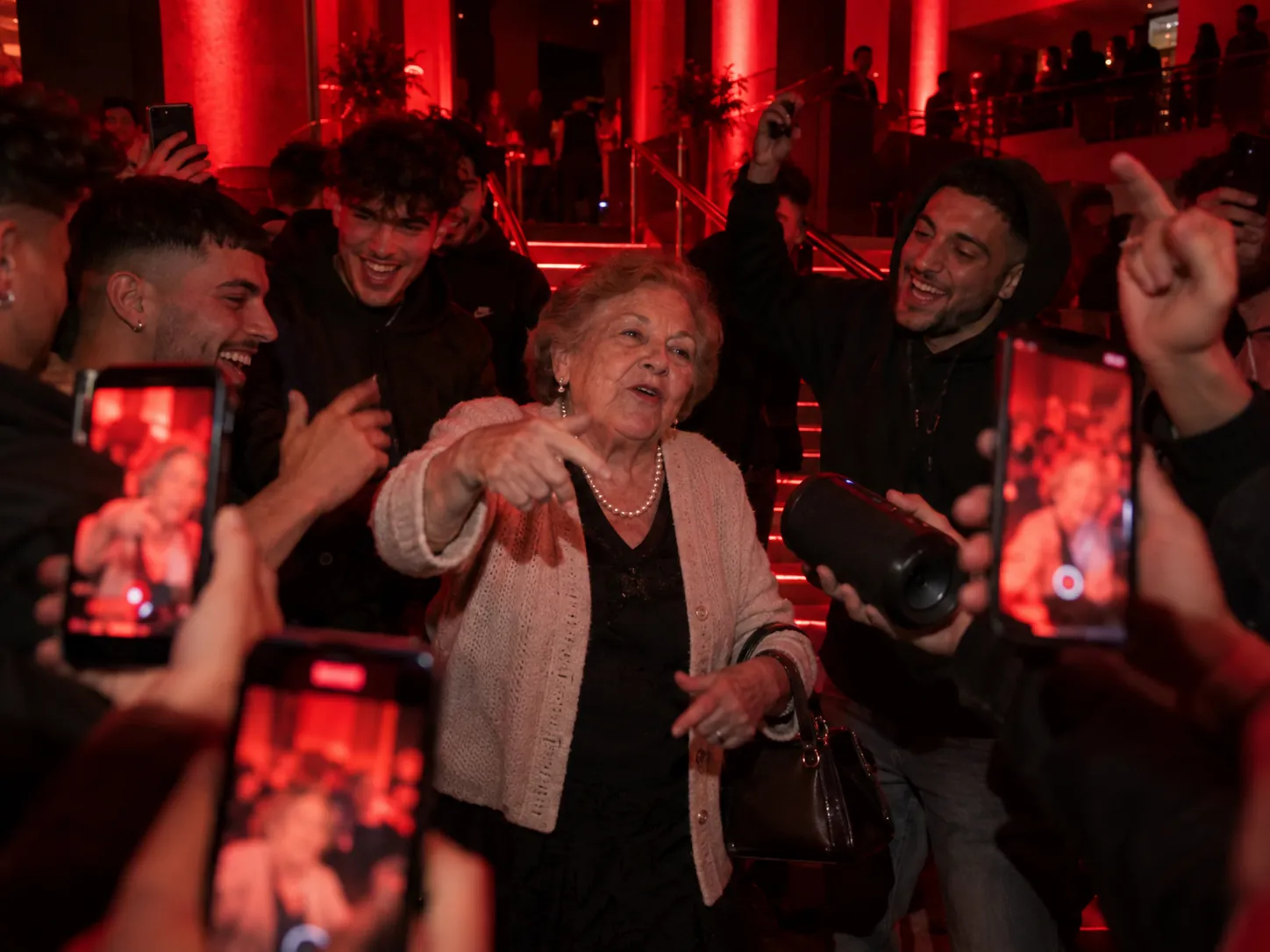 A grandmother in a cardigan and pearl necklace clutching her bag and aiming a finger at a phone camera as she freestyles under red lights, a young crowd around her filming and cheering