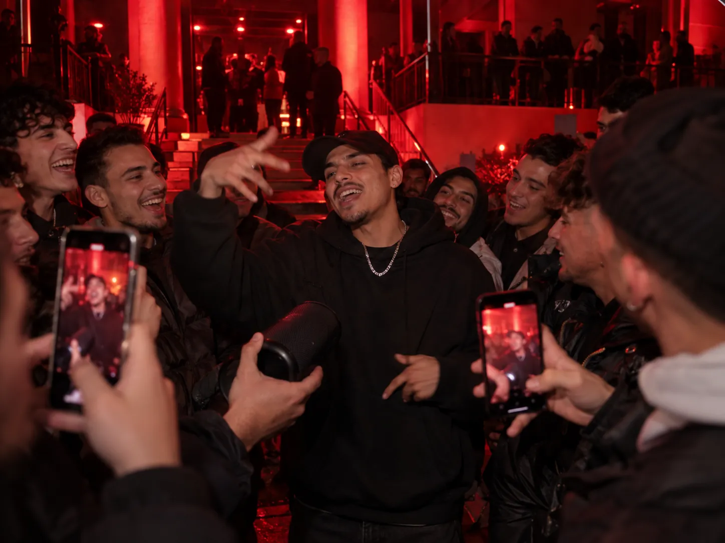 A guy in a cap, silver chain and dark oversized hoodie freestyling under red lights as the people packed around him record on their phones, a black portable speaker held up to him, the Bulin 47 AI trend