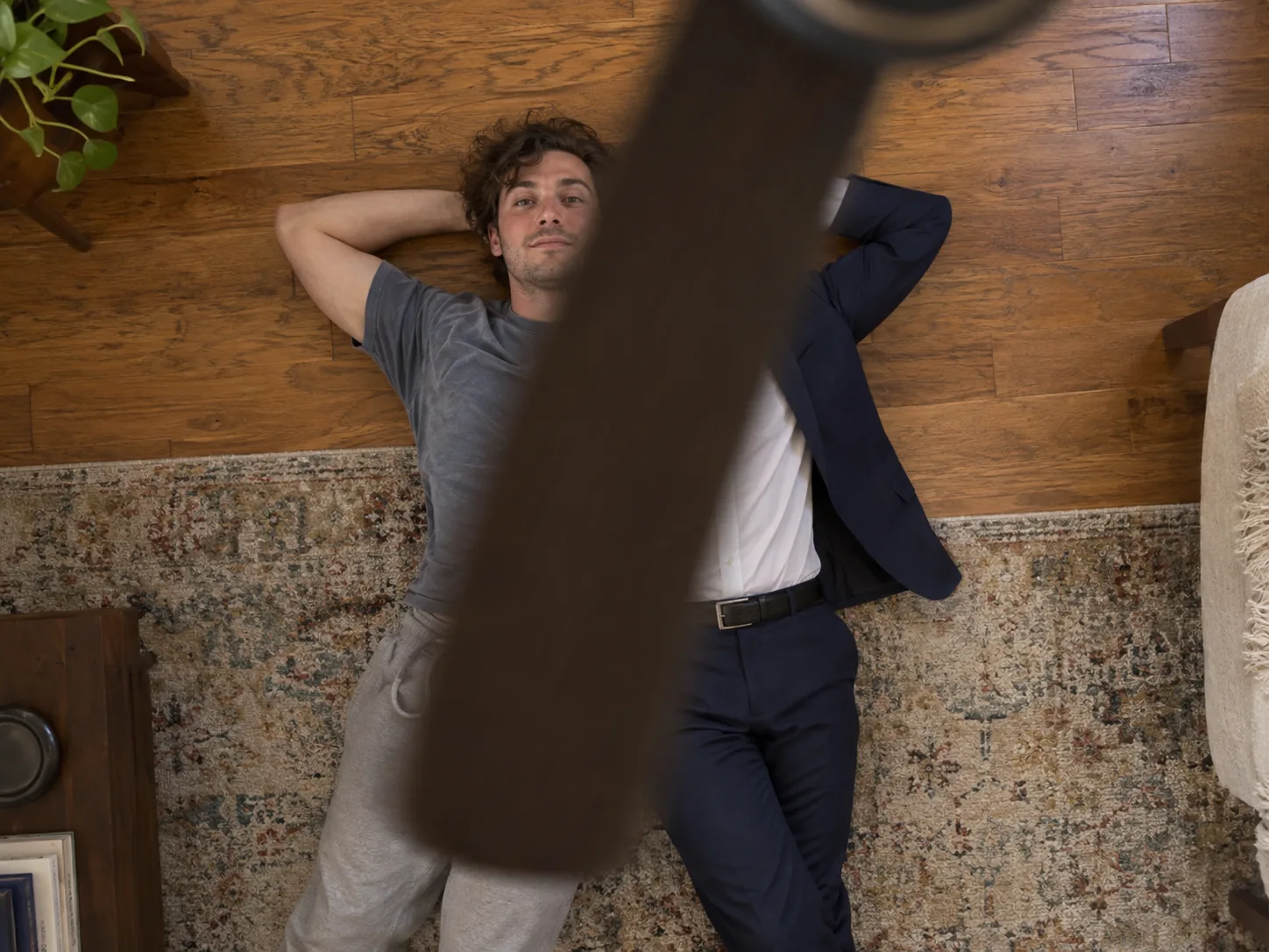 Overhead view past a blurred ceiling fan blade: a man lying on a rug, his outfit changing from a faded T-shirt to a navy suit across the blade