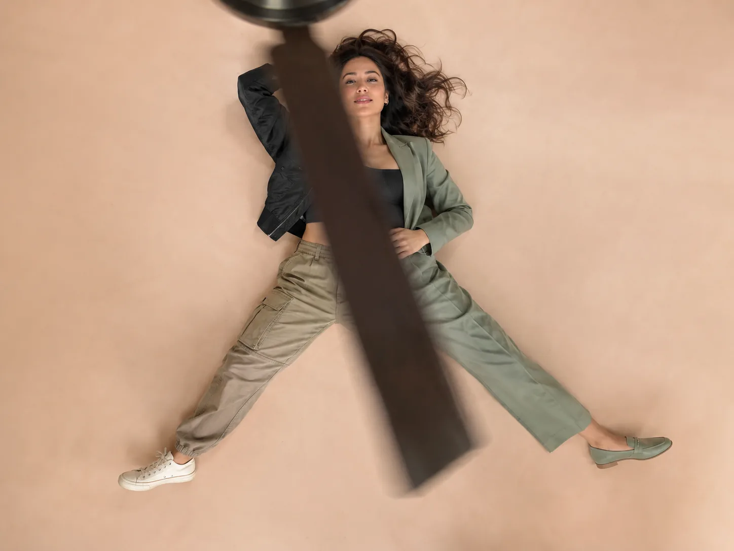 Overhead view past a blurred ceiling fan blade: a woman on a pastel floor, her outfit changing from streetwear to a sage-green suit across the blade
