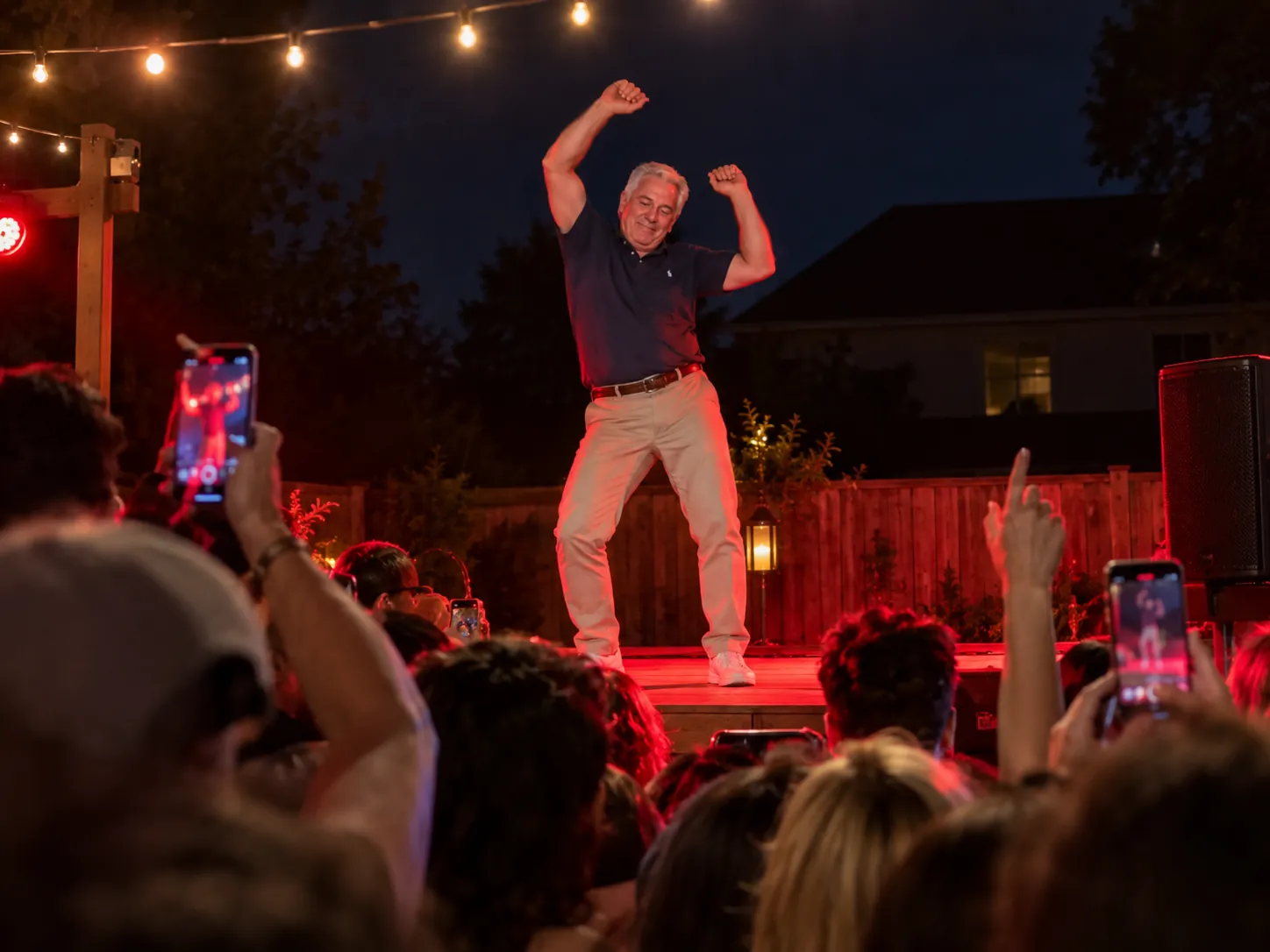 A grey-haired man in a polo shirt dancing with his hands thrown overhead on a red-lit outdoor stage, phones raised in front of him