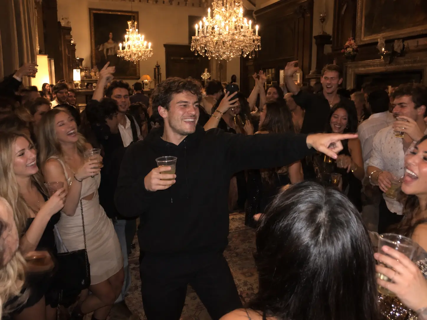 A young man in a black hoodie holding a party cup and pointing into a cheering crowd under crystal chandeliers at a house party