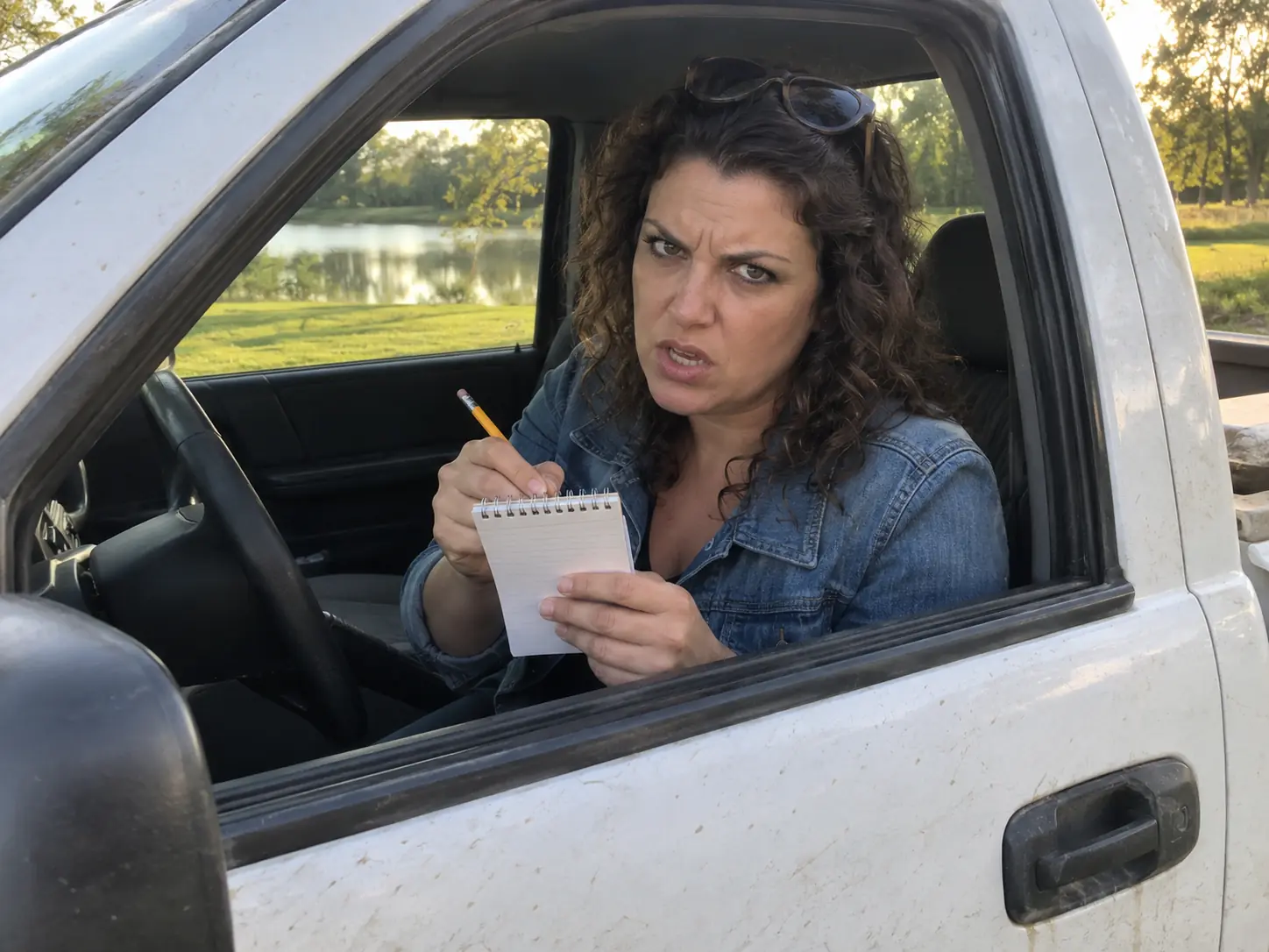 A woman with curly hair in a denim jacket holding up a notepad at the window of a white pickup truck, ready to take someone's name