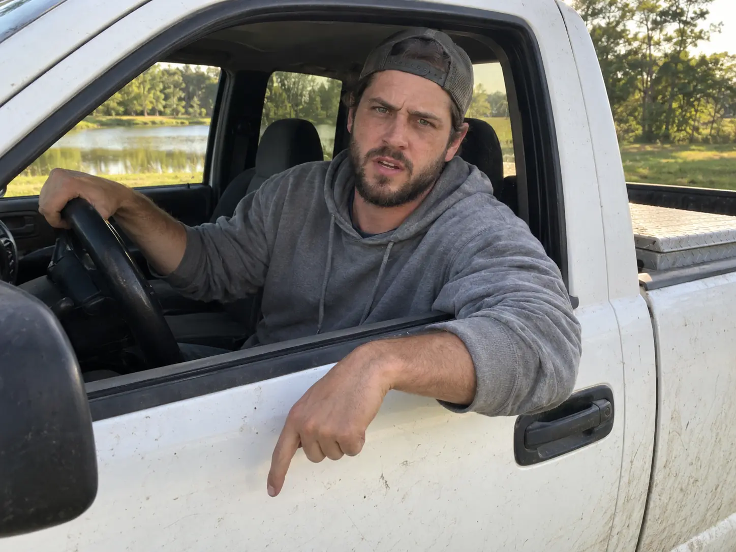 A bearded man in a backwards cap glaring out of the driver's window of a white pickup truck by a pond, the Hit the Road Dude meme