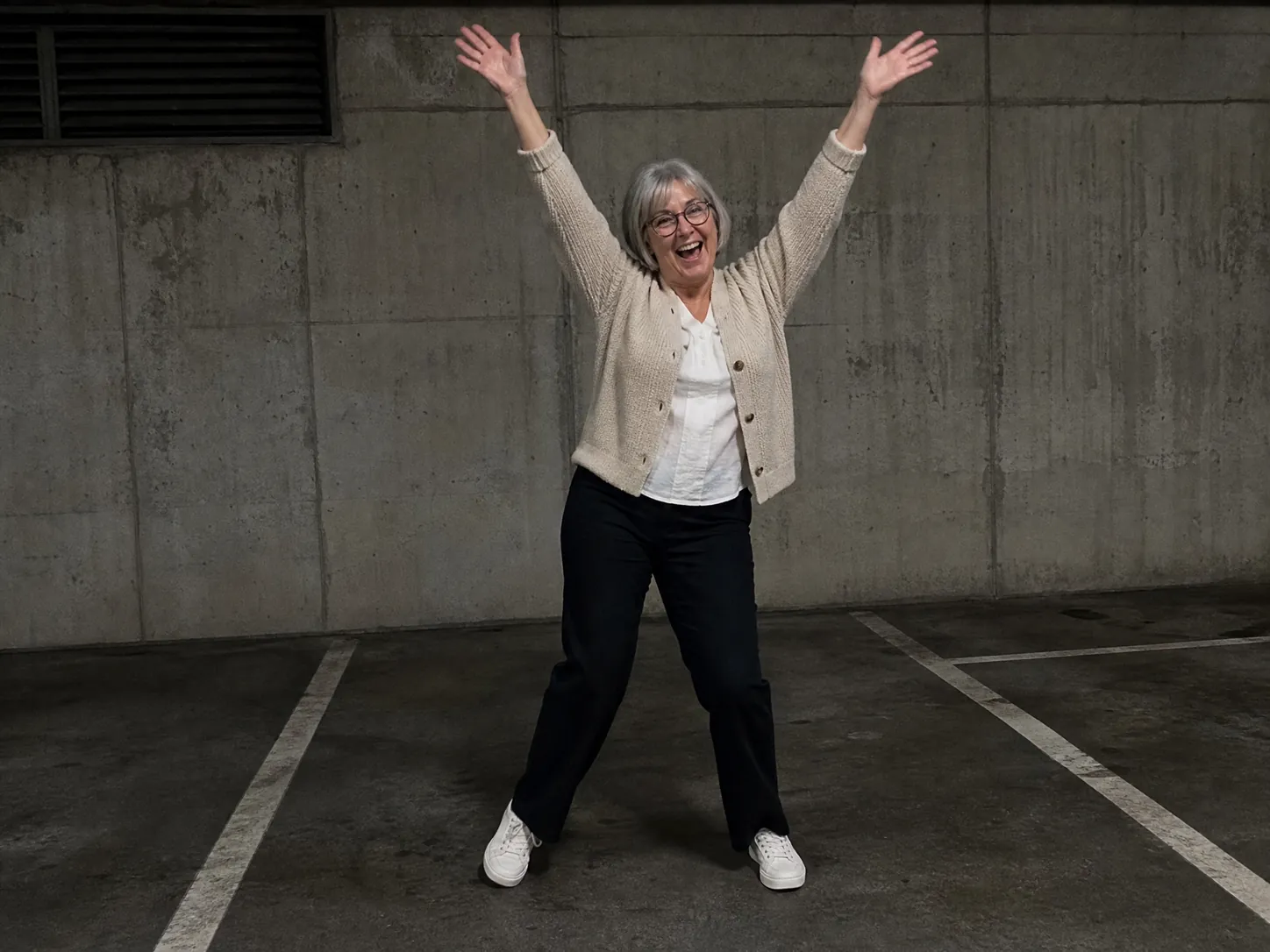 A grey-haired mum in glasses and a beige cardigan throwing both arms overhead and laughing in front of a concrete wall, a family Lisa SaWaDiKa dance video