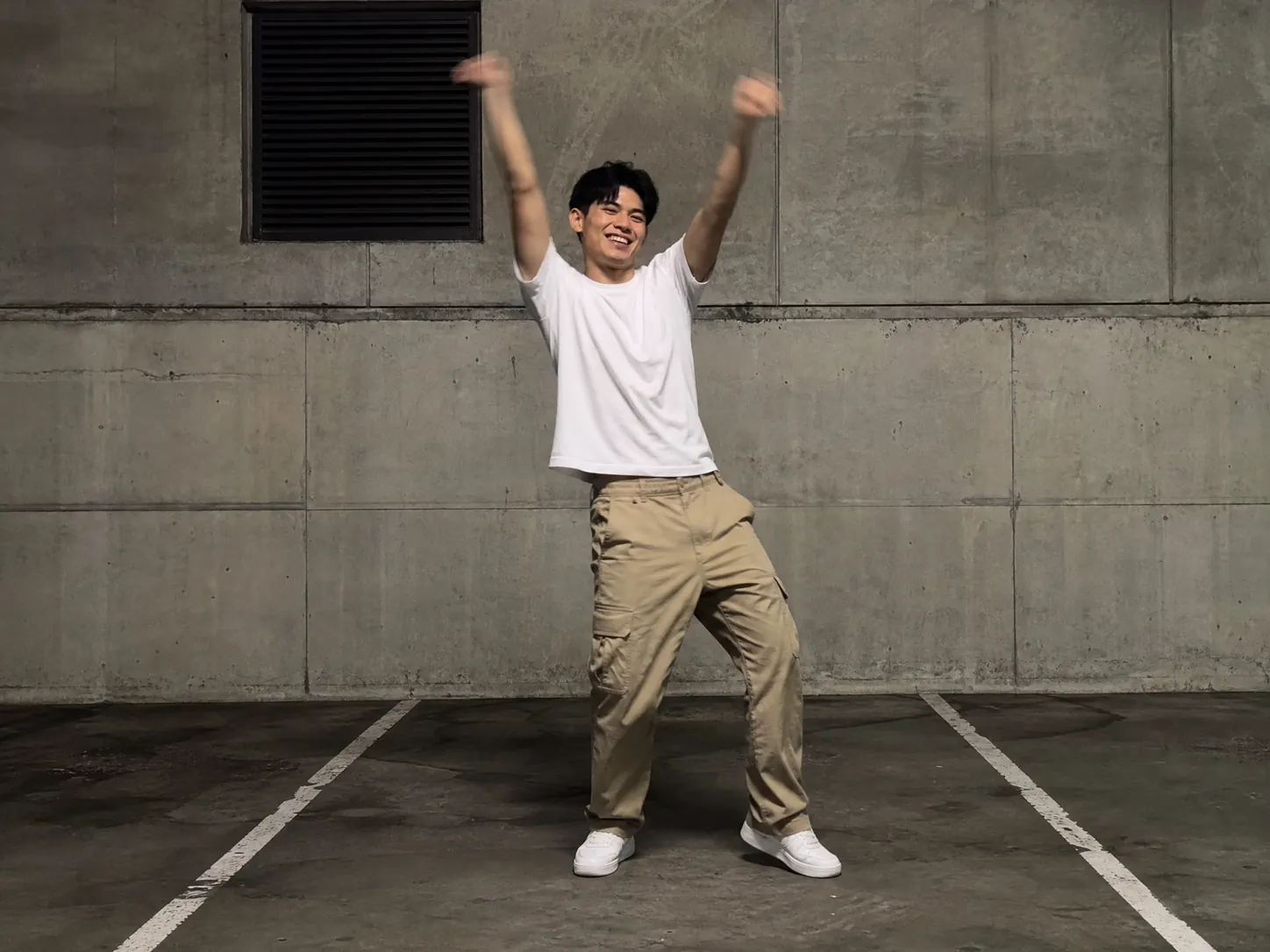 A grinning guy in a plain white tee and khaki cargo pants throwing both arms overhead in the SaWaDiKa dance against a concrete wall, a friend's Lisa SaWaDiKa dance AI video