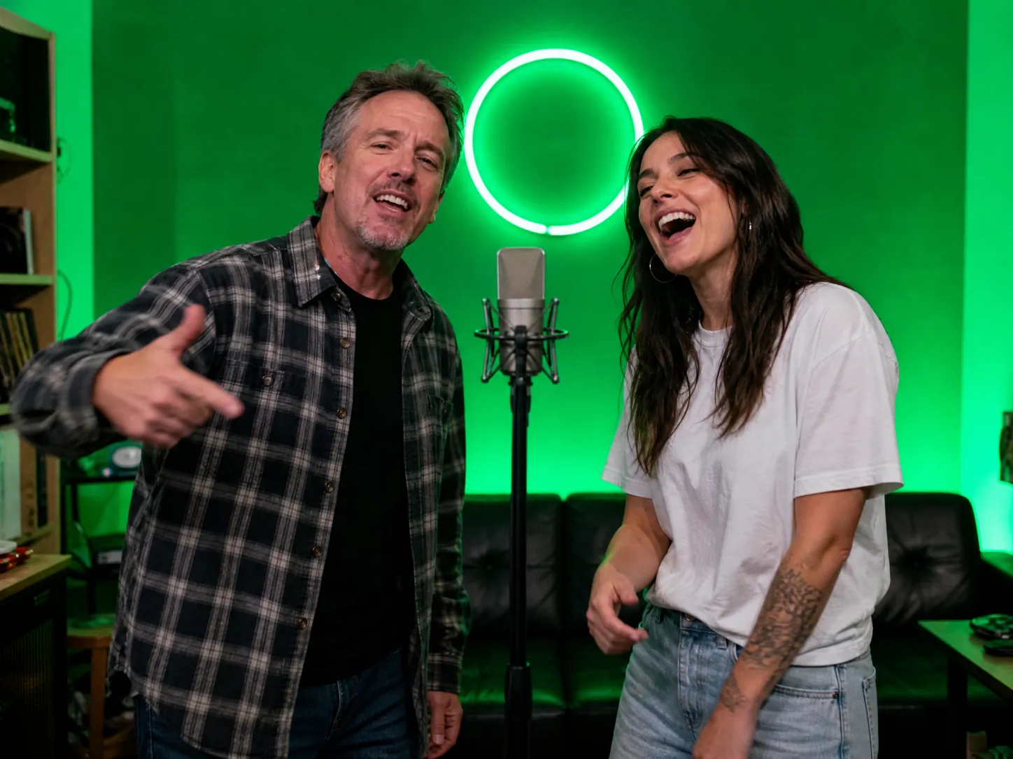 A father and his adult daughter grinning as they perform side by side at a microphone in front of a green wall