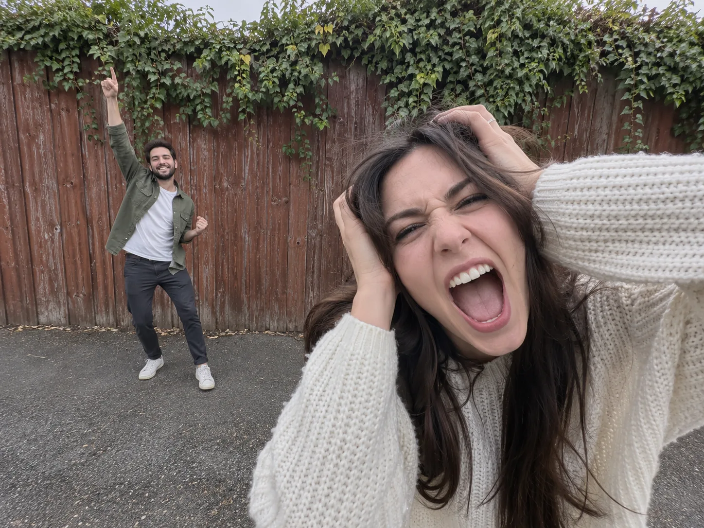 A woman in a cream sweater gripping her hair and screaming into the lens while her boyfriend in a green overshirt dances behind her by an ivy-covered fence
