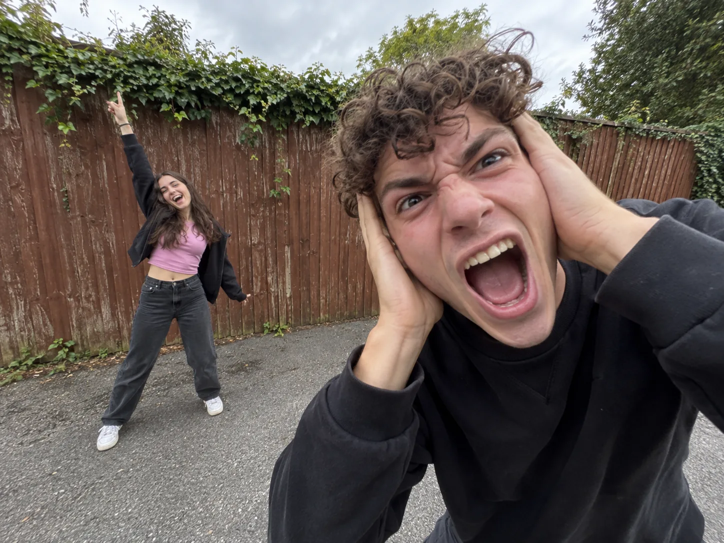 A curly-haired young man clutching his head and screaming close to an ultra-wide lens while a young woman in pink and black dances behind him by a red wooden fence, the Smalltown Boy AI trend