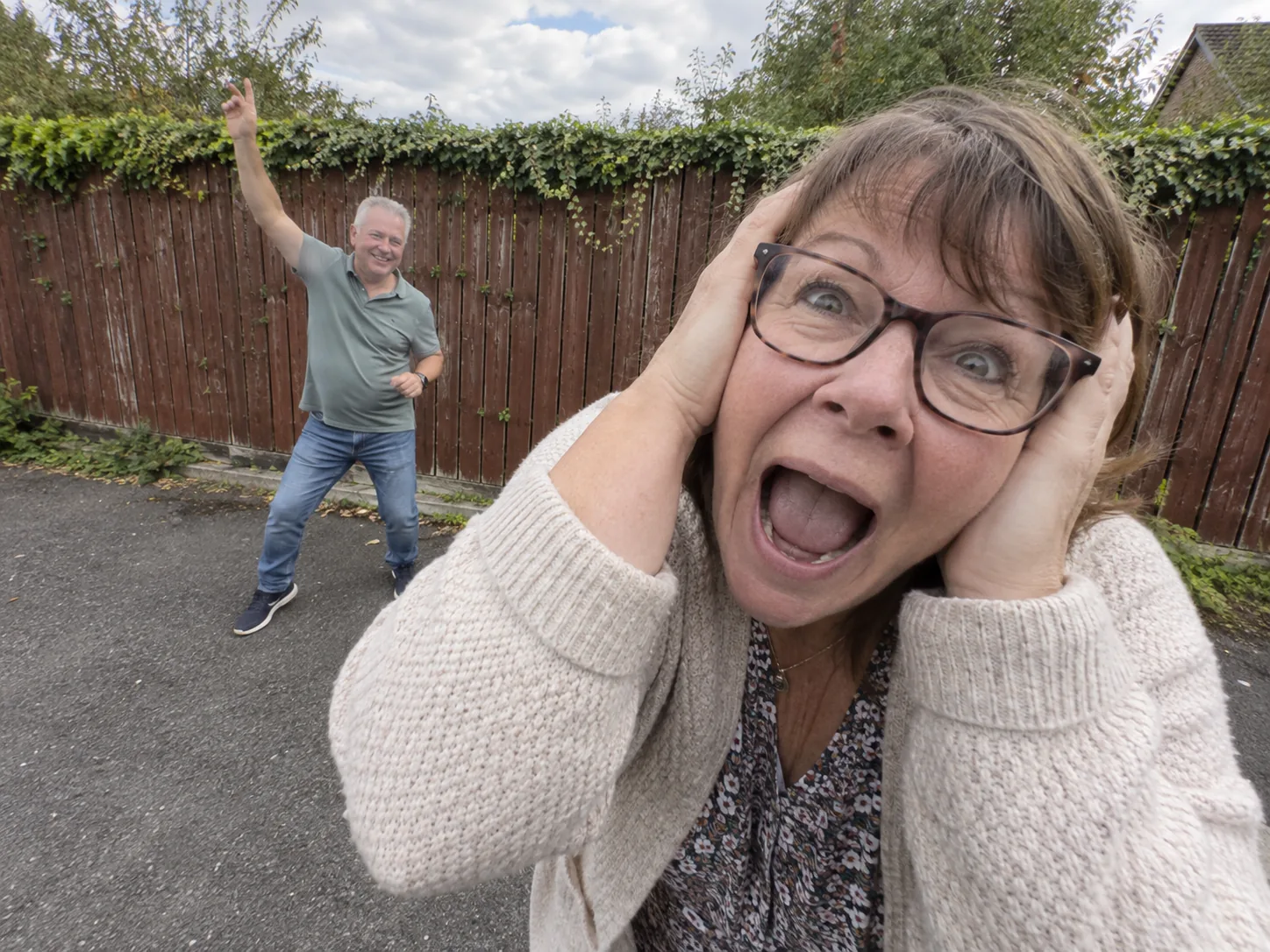 A mum in glasses and a cardigan screaming with her hands on her head close to the lens while a grey-haired dad dances behind her with one arm up, a parents version of the Smalltown Boy dance