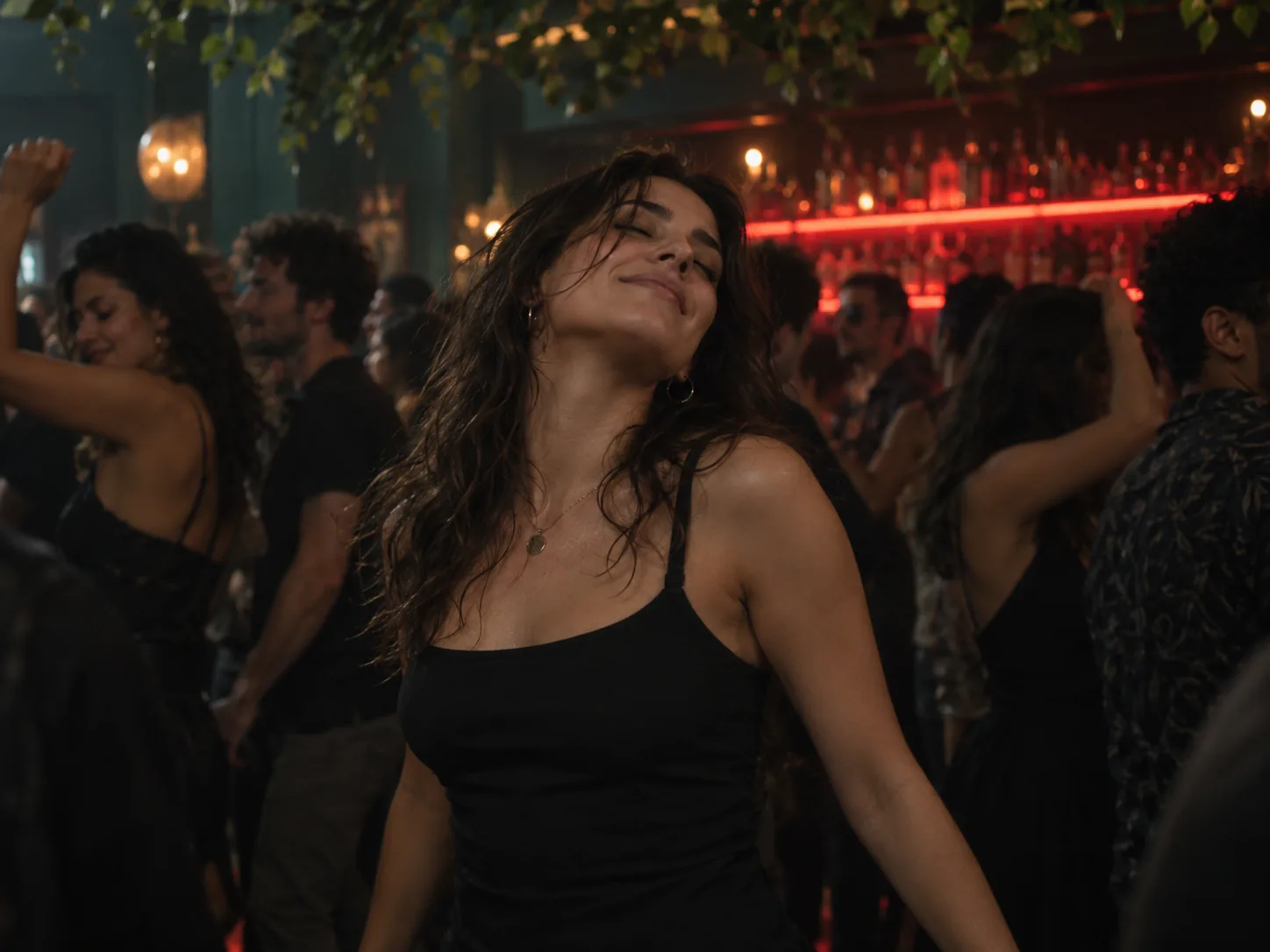 A young woman with her eyes closed and chin up, dancing in a packed nightclub under a red neon bar