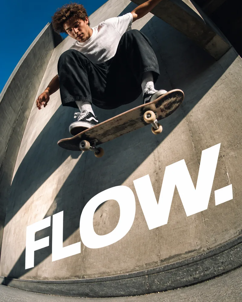 A skateboarder frozen at the peak of a trick above curved concrete, shot on a low fisheye lens with the word FLOW painted across the wall
